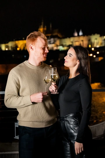Couple toasting with wine on the terrace at night with Prague Castle lit up.