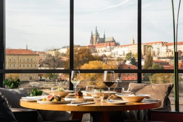Prostřený stůl s pokrmy a vínem u panoramatického okna s výhledem na Pražský hrad.
